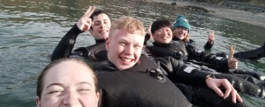 Six divers in wetsuits are standing in shallow water smiling at the camera